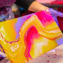 Person holding abstract painting with vibrant pink, yellow, and purple swirls, wearing gloves.