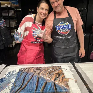 Two people in aprons smiling, posing with painted canvas.