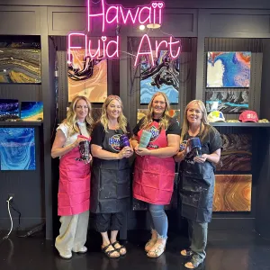 Four women in aprons hold art pieces, standing under a 'Hawaii Fluid Art' neon sign.