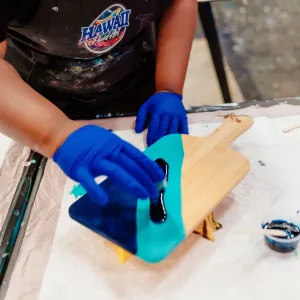 Person painting wooden board with blue resin, wearing gloves.
