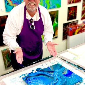 Man in purple apron displays a blue abstract painting in art studio.